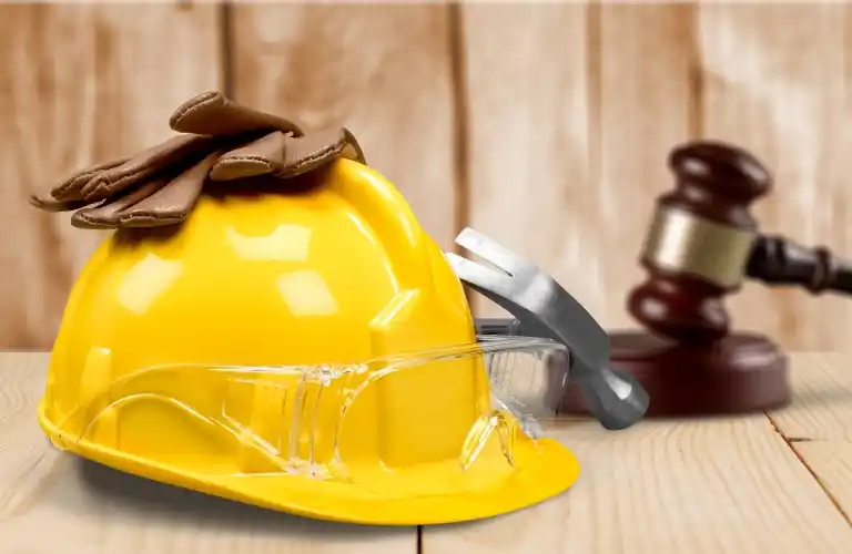 Yellow construction helmet with safety gloves and glasses, hammer and wooden gavel in background.