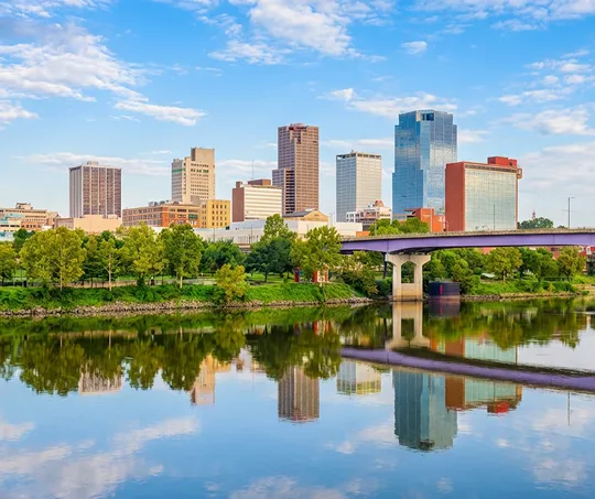 City skyline with modern buildings, a bridge, green trees, and river reflections under a partly cloudy sky.