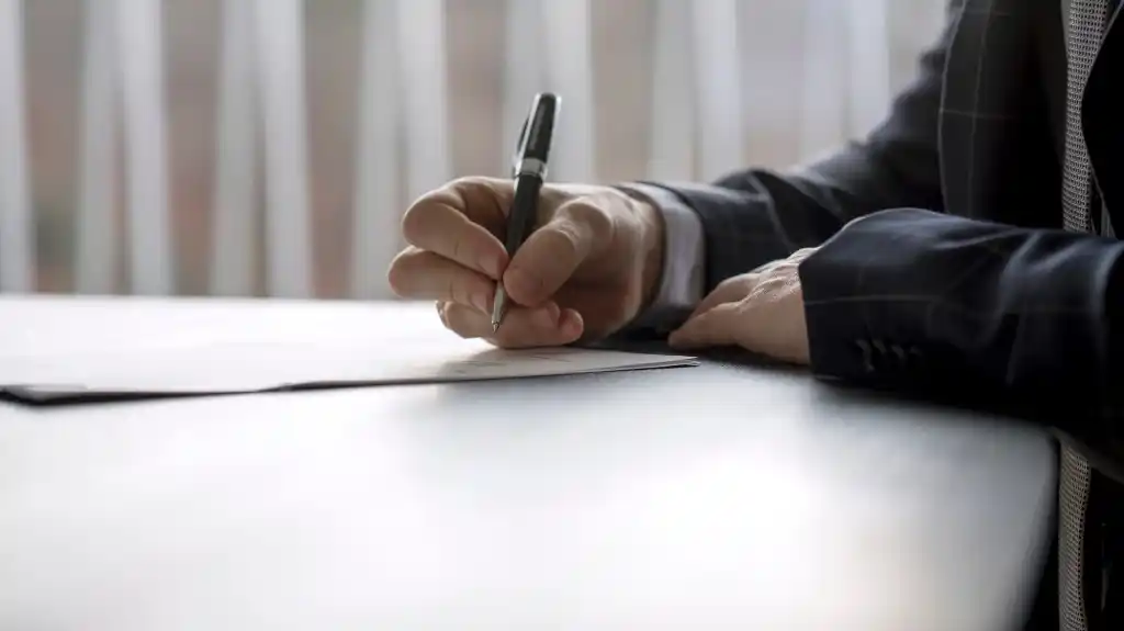 Close-up of a person in a dark suit writing on white paper with a black pen.
