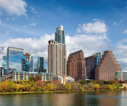 City skyline with modern high-rise buildings and autumn foliage along a waterfront under a partly cloudy sky.