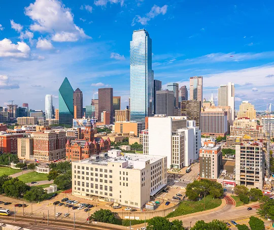 A clear daytime cityscape of downtown Dallas, Texas, featuring modern skyscrapers, historic buildings, and a bright blue sky with scattered clouds.