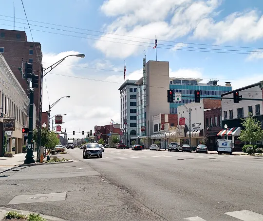 Urban street scene with commercial buildings, vehicles, and traffic lights under a partly cloudy sky.