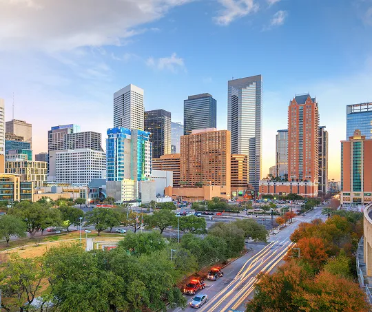 City skyline at dusk with modern high-rise buildings, green trees, and light trails on the road.