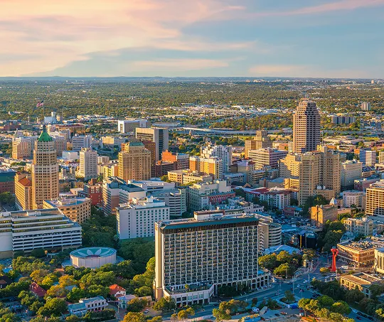 Aerial view of a cityscape with diverse high-rise buildings, green trees, and a partly cloudy sky at sunset.