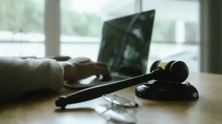 Wooden judge's gavel on desk with blurred laptop, eyeglasses, and person typing in background.