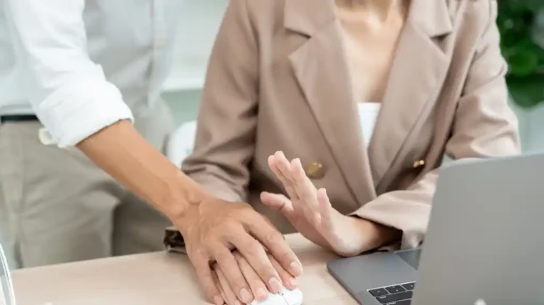 Woman in beige blazer rejecting man's hand over desk with laptop in office setting
