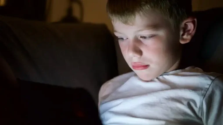 Young boy in white shirt illuminated by screen light, focused in a dark room.