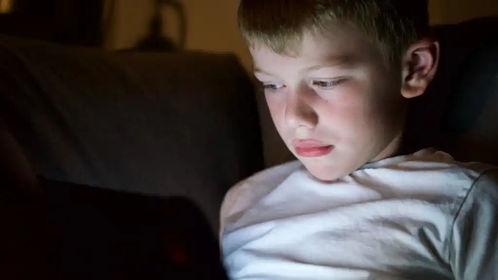 Young boy in white shirt illuminated by screen light, focused in a dark room.