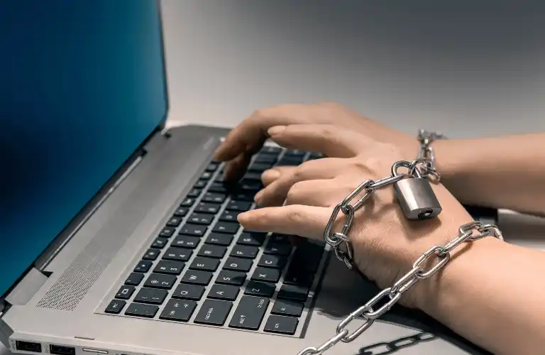 Hands typing on a laptop keyboard, bound by a metal chain and padlock.