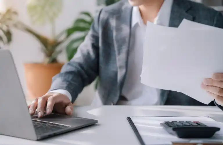 Professional person in gray blazer working on laptop and holding papers at desk.