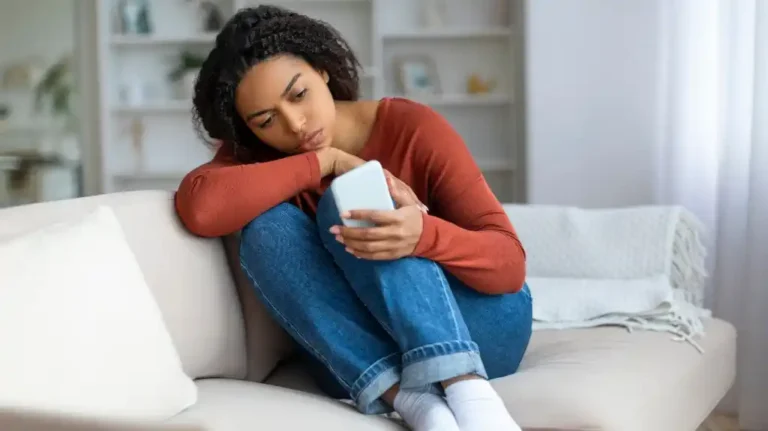 Young woman in rust-colored sweater and jeans sitting on beige sofa, looking at smartphone.