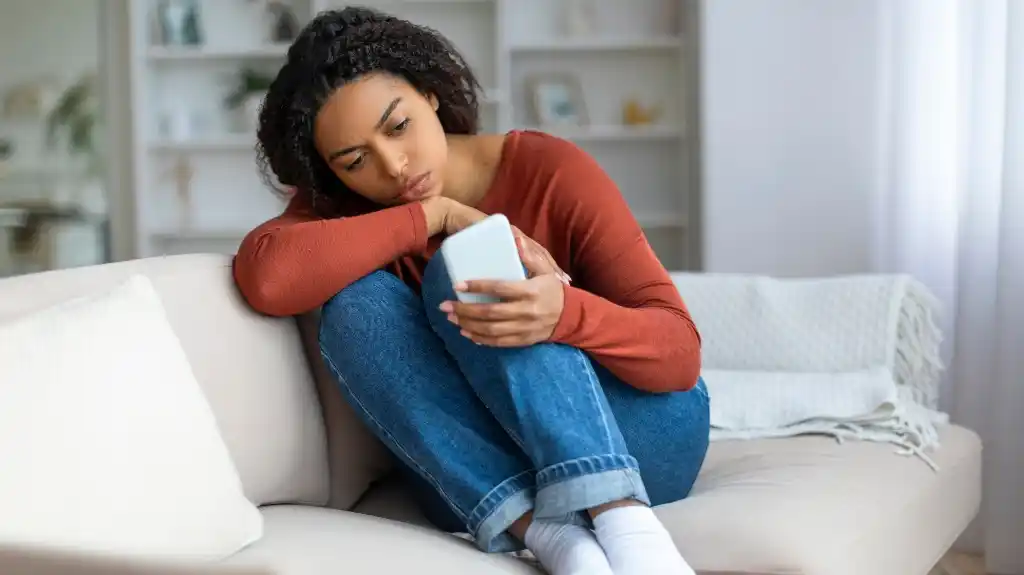 Young woman in rust-colored sweater and jeans sitting on beige sofa, looking at smartphone.