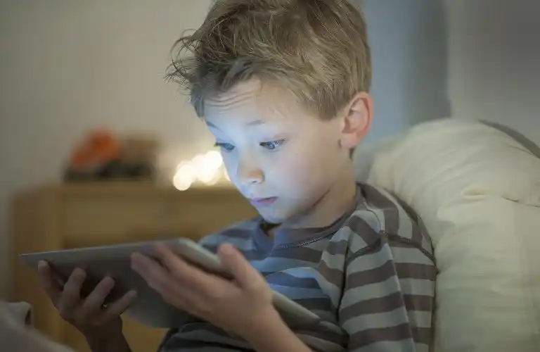 Young boy in striped shirt intently looking at a tablet screen in dimly lit room.