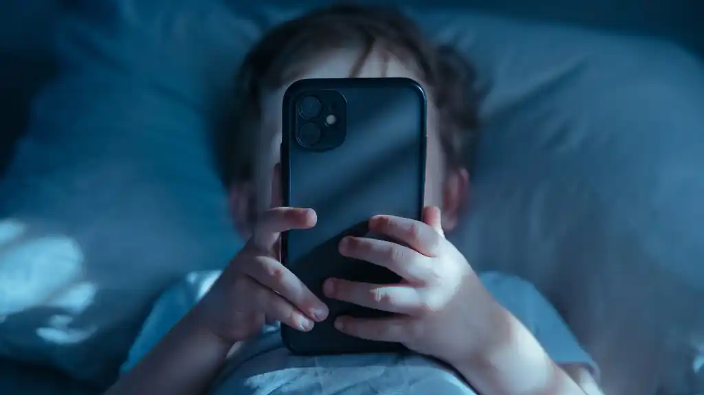 Child lying in bed holding a smartphone close to their face in dim lighting.